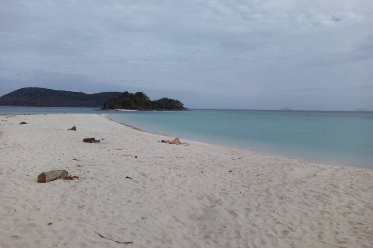 Ein Strand mit weißem Sand und ruhigem Wasser, eine kleine Insel in der Ferne und ein bewölkter Himmel.