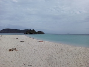 Una playa de arena blanca y agua tranquila, una pequeña isla a lo lejos, y un cielo nublado.