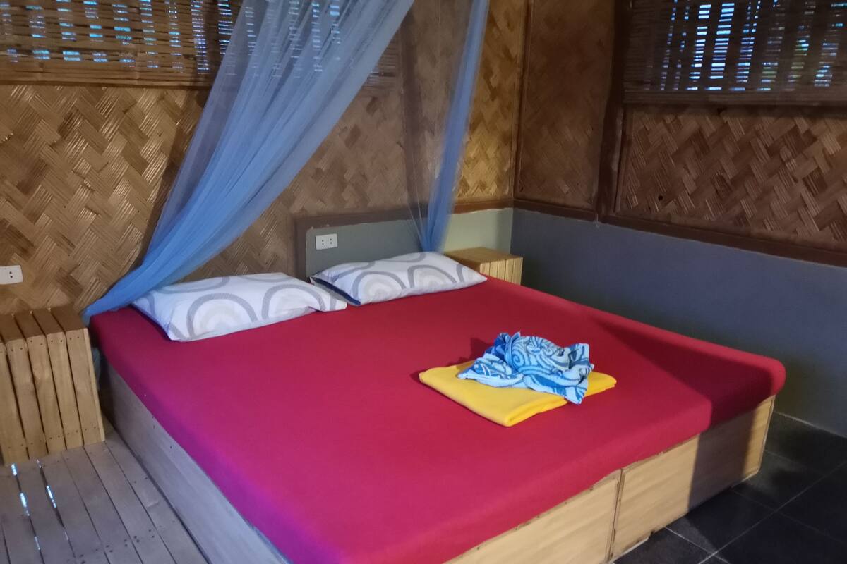 Ein Bett mit roter Bettdecke, zwei Kissen und einem blau-weißen Stoffüberwurf.