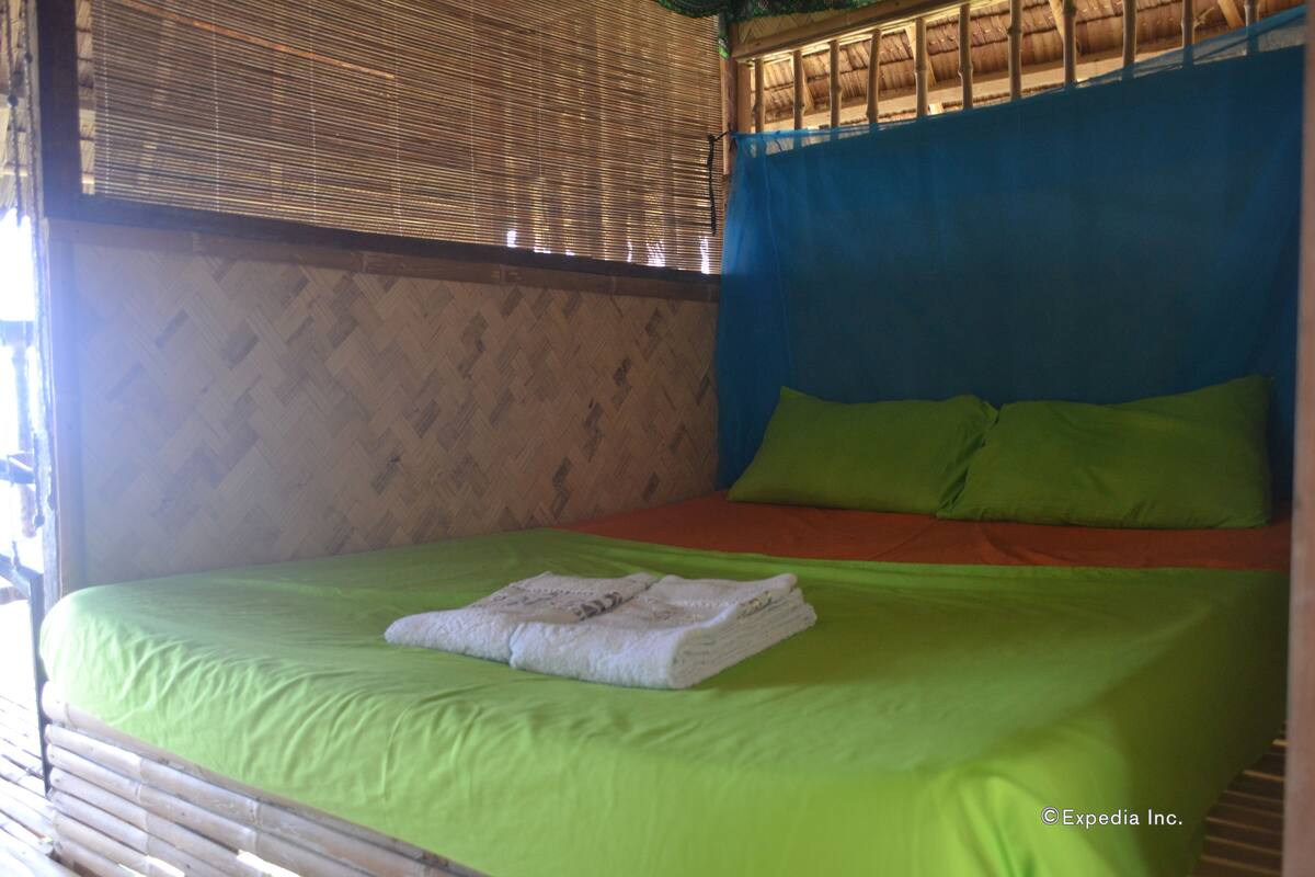 Ein Zimmer mit einem Bett, grüner Bettwäsche, einem gefalteten weißen Handtuch und einem blauen Vorhang.