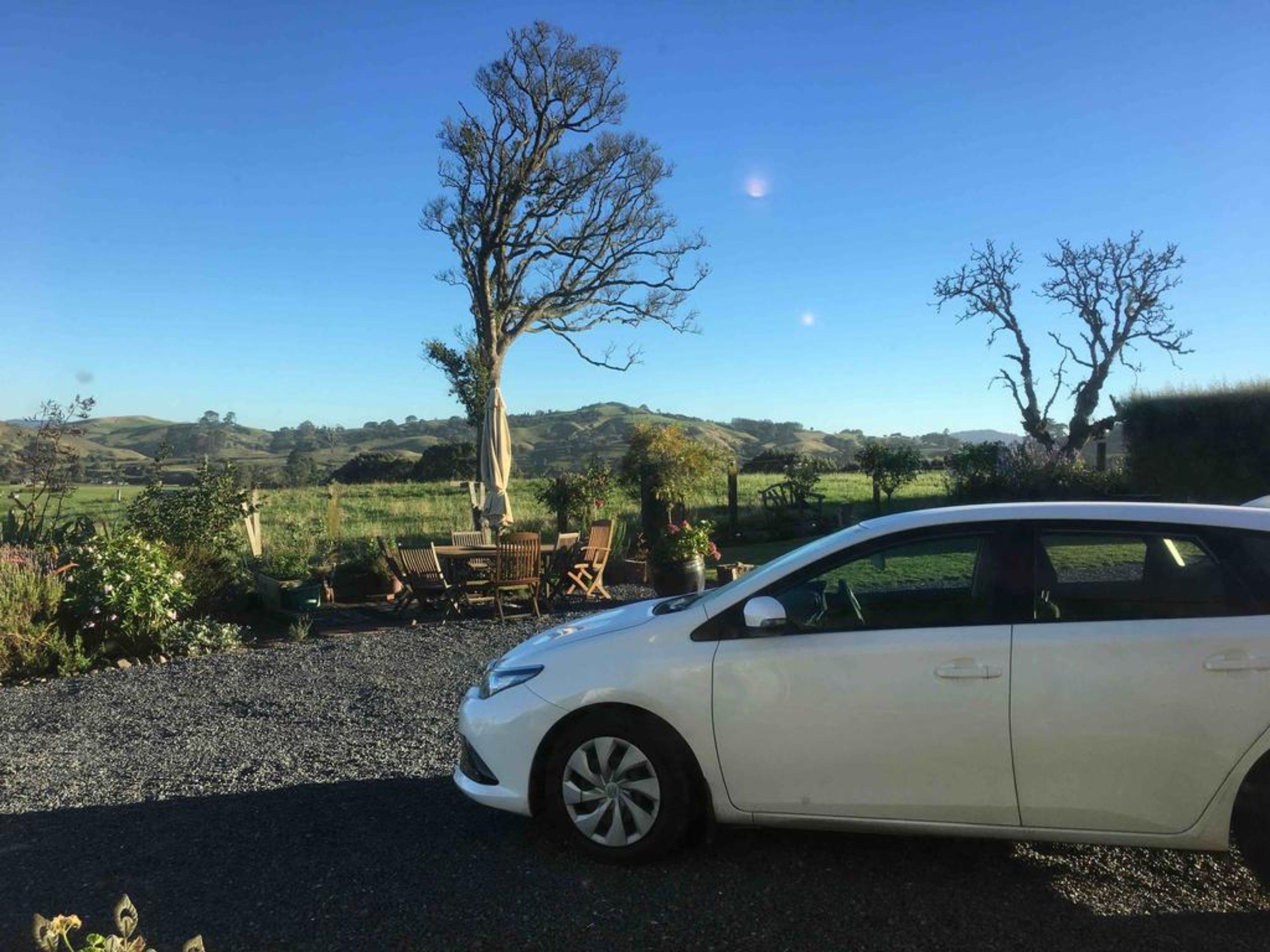 Un coche blanco estacionado en un entorno rural, con un camino de grava, una mesa y sillas de madera, y un árbol grande al fondo.