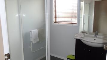 Un baño con cabina de ducha de vidrio, lavamanos blanco y ventana con persianas.