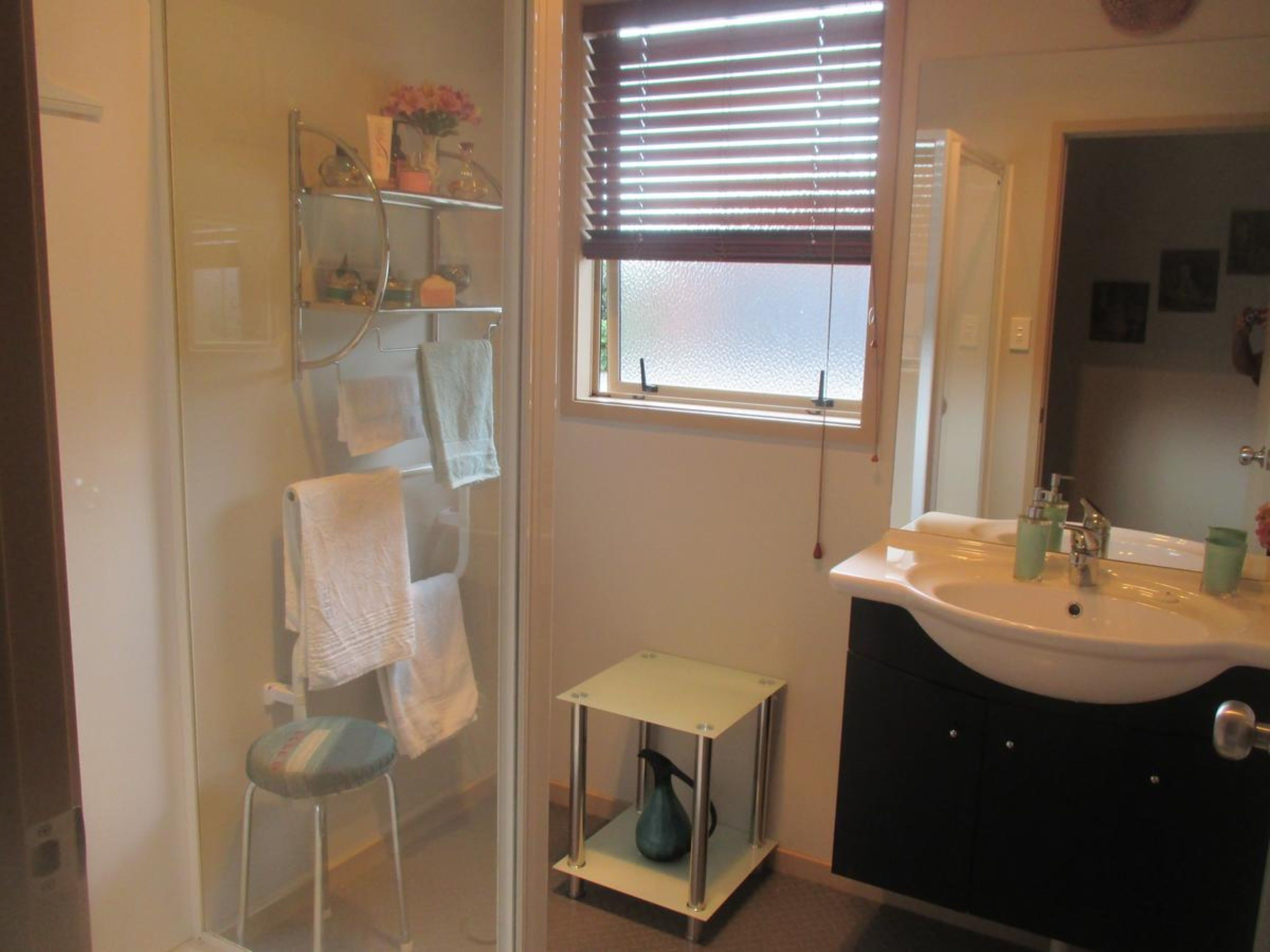 A bathroom with a glass shower enclosure, a small stool, a window with blinds, a sink with a mirror, and a shelf with toiletries.