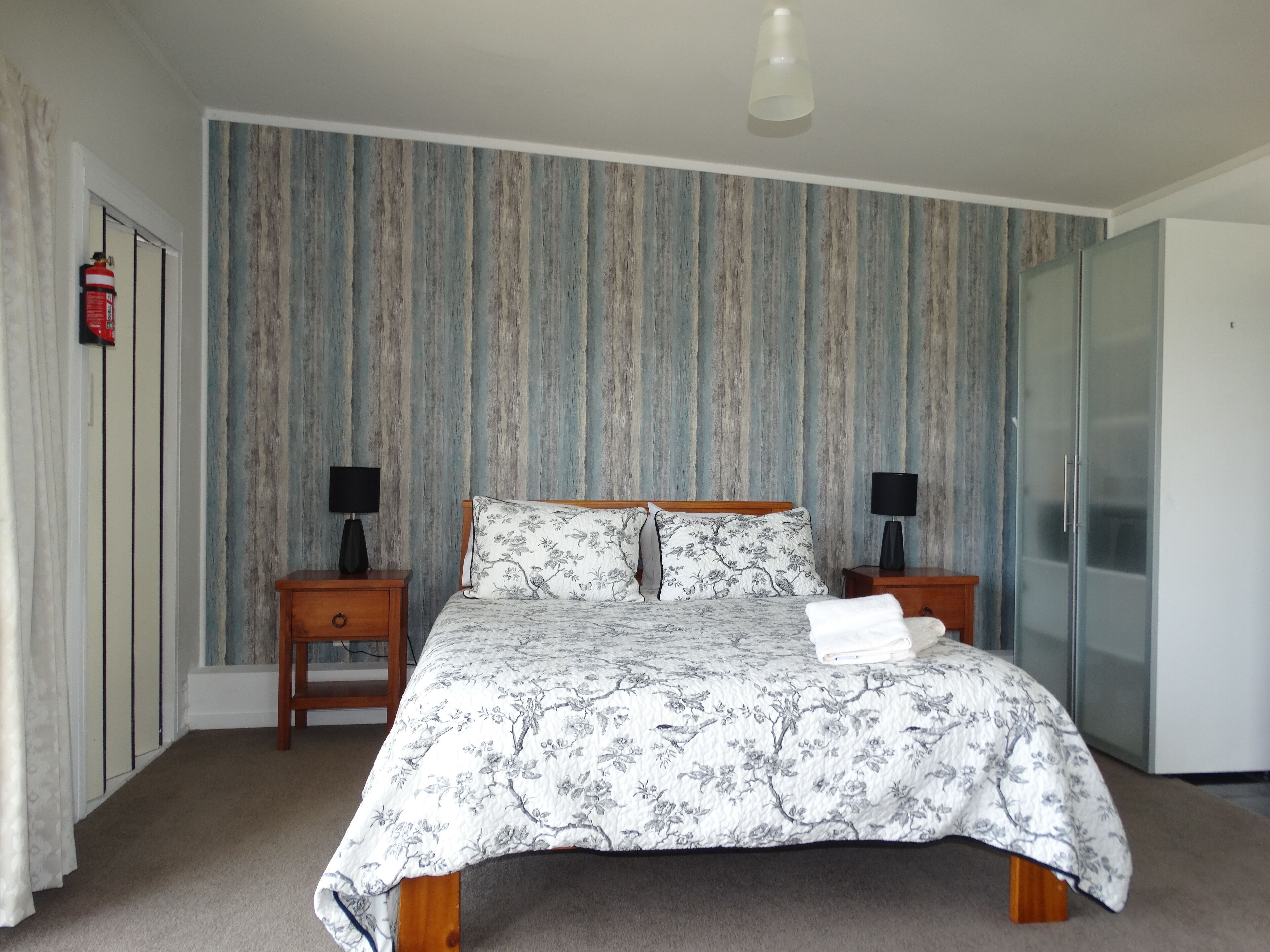 A bedroom with a bed, two bedside tables with lamps, a wardrobe, and a door with a fire alarm.