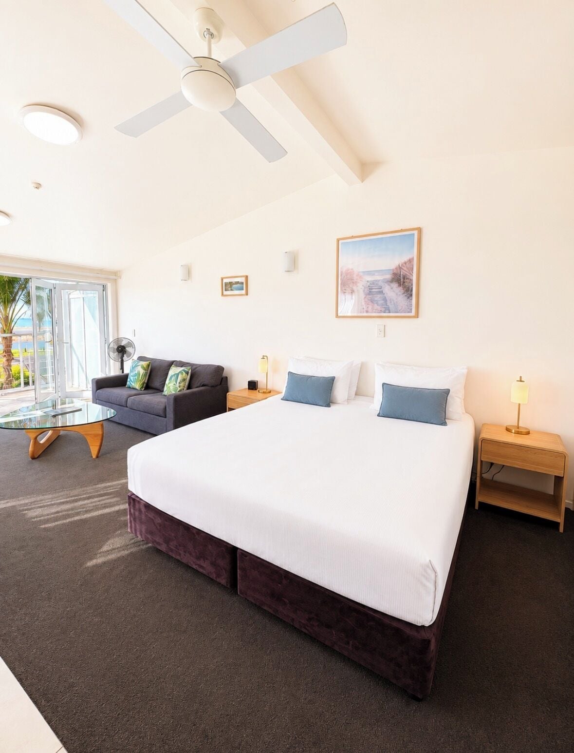 A hotel room with a large bed, a sofa, a glass coffee table, and a ceiling fan.