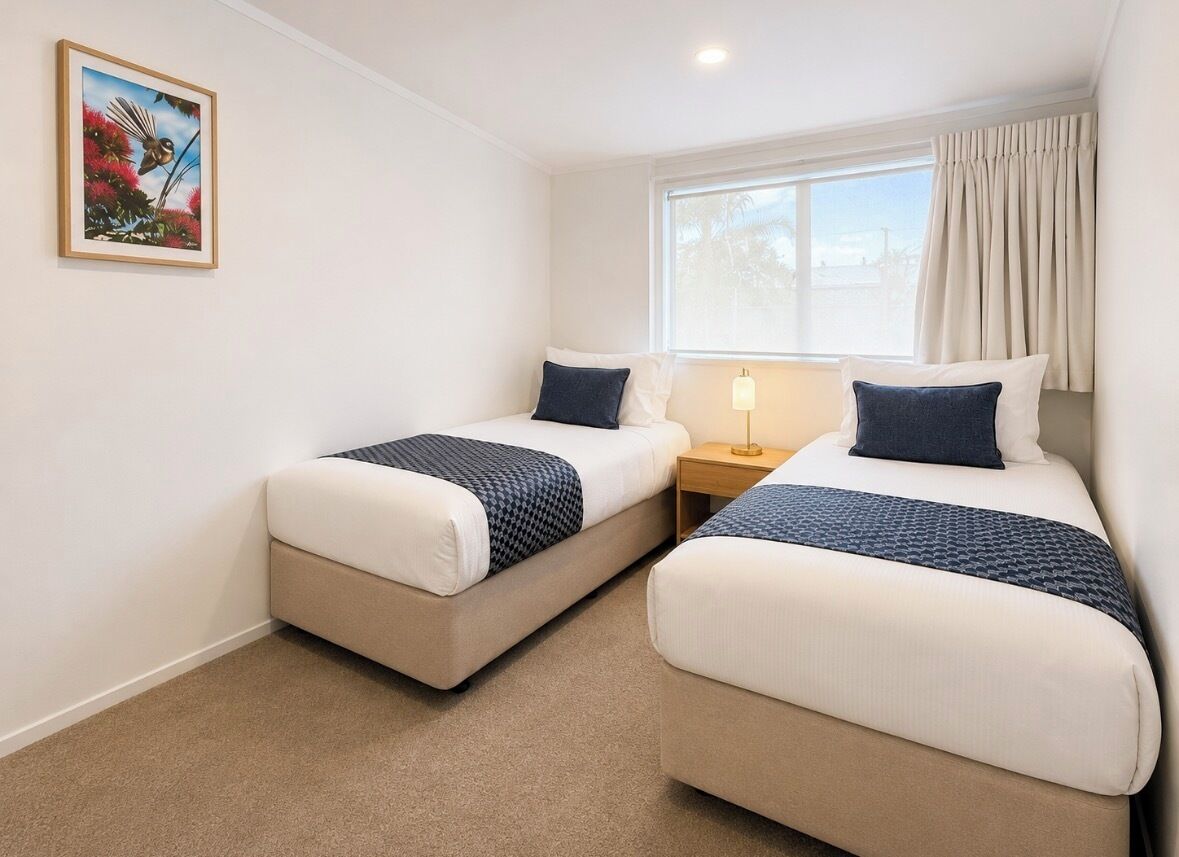 A room with two beds, a nightstand, a lamp, a window with curtains, and a framed picture on the wall.