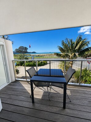 En balkon med et bord og stole, der giver udsigt over et kystlandskab med palmer og en klar himmel.