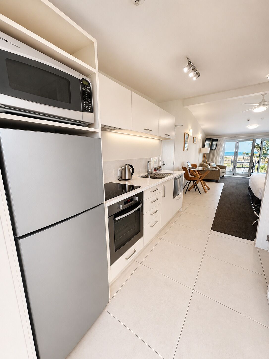 A modern kitchen with a built-in microwave, oven, and stovetop. There is a dining area with a table and chairs, and a large window offering a view of the beach.
