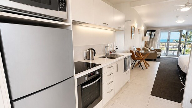 Une cuisine moderne équipée d’un four à micro-ondes, d’un four et d’une plaque de cuisson intégrés. On y trouve également un coin repas avec une table et des chaises, ainsi qu’une grande fenêtre offrant une vue sur la plage.