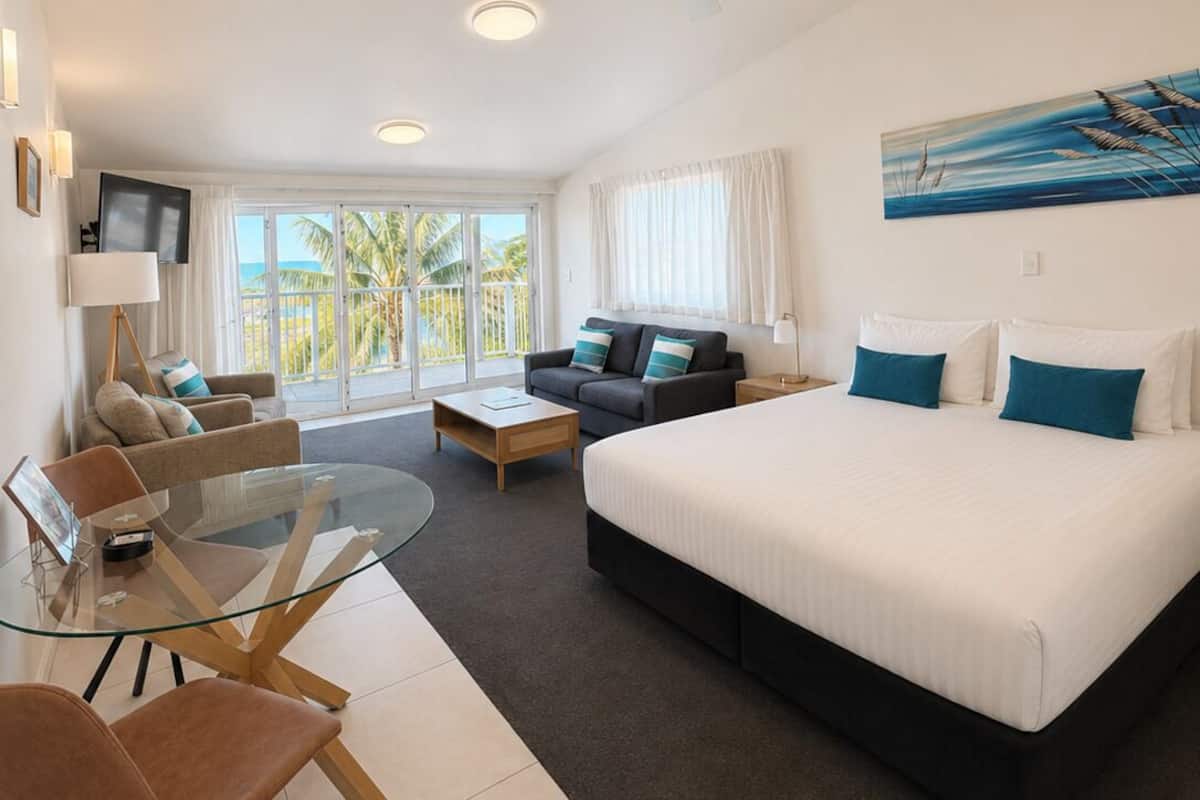 A modern hotel room with a large bed, a sofa, a glass dining table, and a ceiling fan.