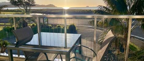 A balcony with a table and chairs, overlooking a body of water and a distant tree line.