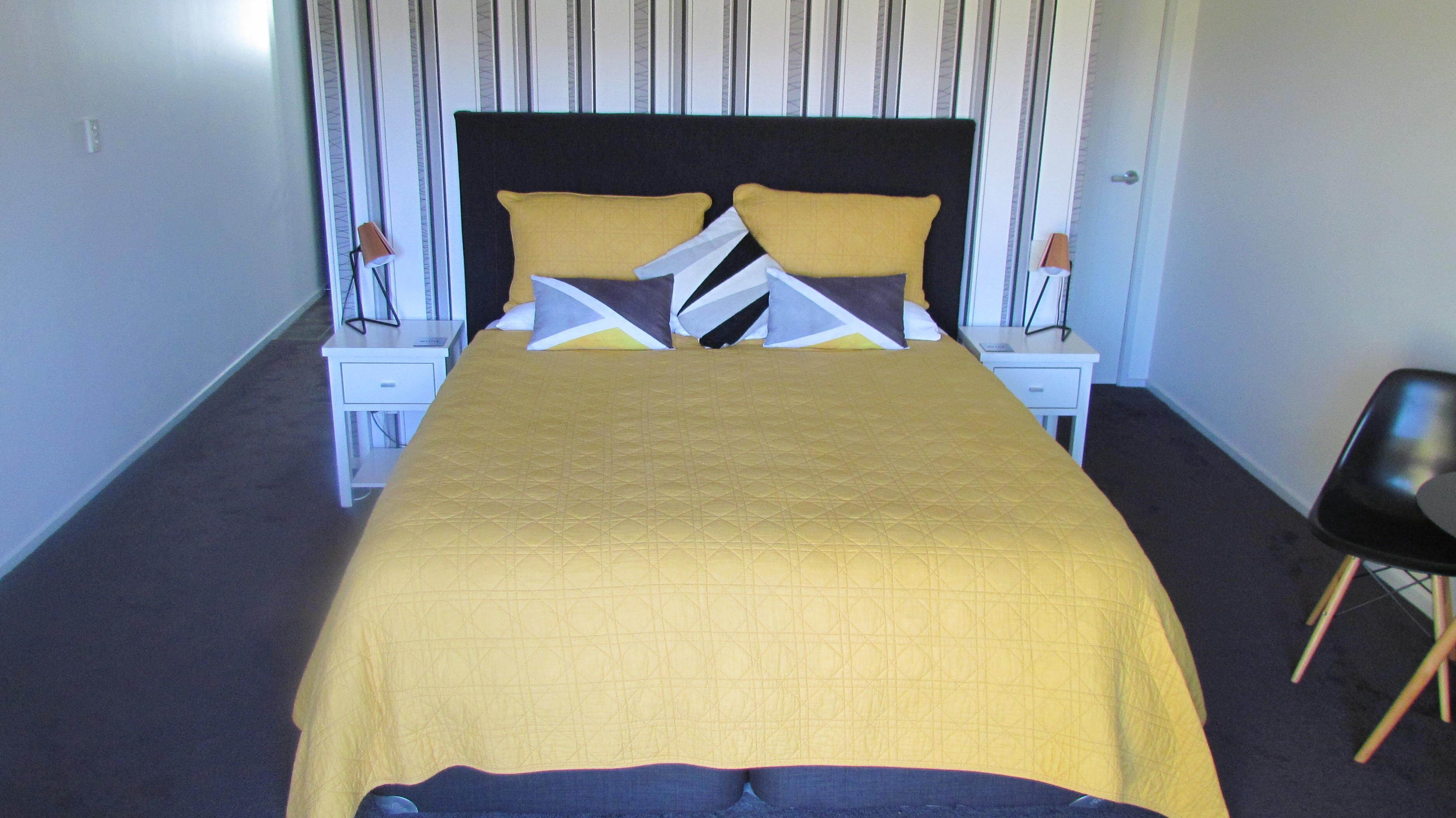 Un lit bien fait, recouvert d’une couvre-lit jaune et agrémenté d’oreillers aux motifs géométriques, flanqué de deux tables de chevet blanches surmontées de lampes, dans une pièce dont le papier peint est rayé et qui contient une chaise noire.