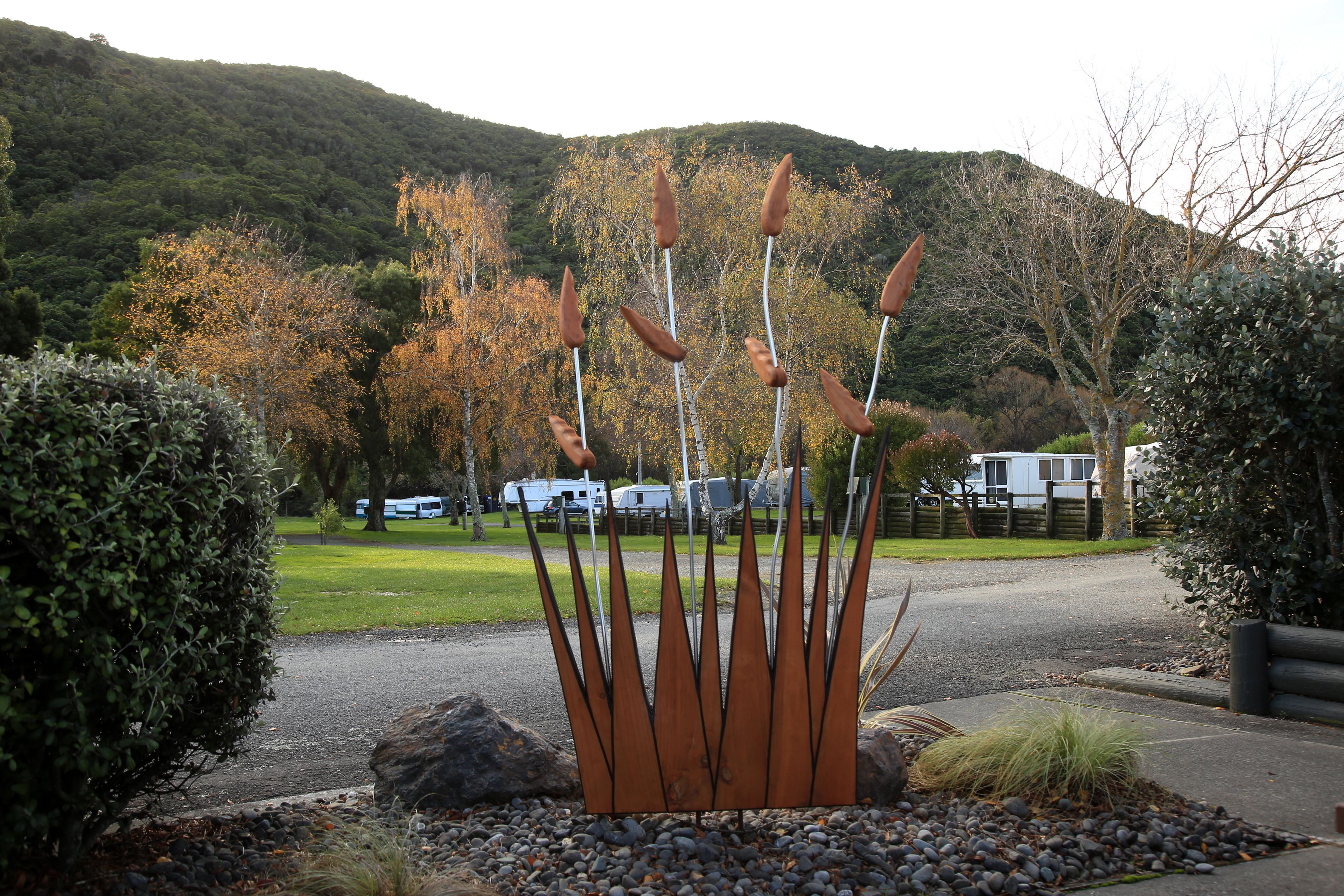 A metal sculpture with multiple pointed elements in a landscaped area with rocks and shrubs, set against a backdrop of trees and a hill.