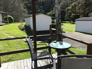 A wooden deck with a table and chairs, overlooking a well-maintained lawn and a small white building.