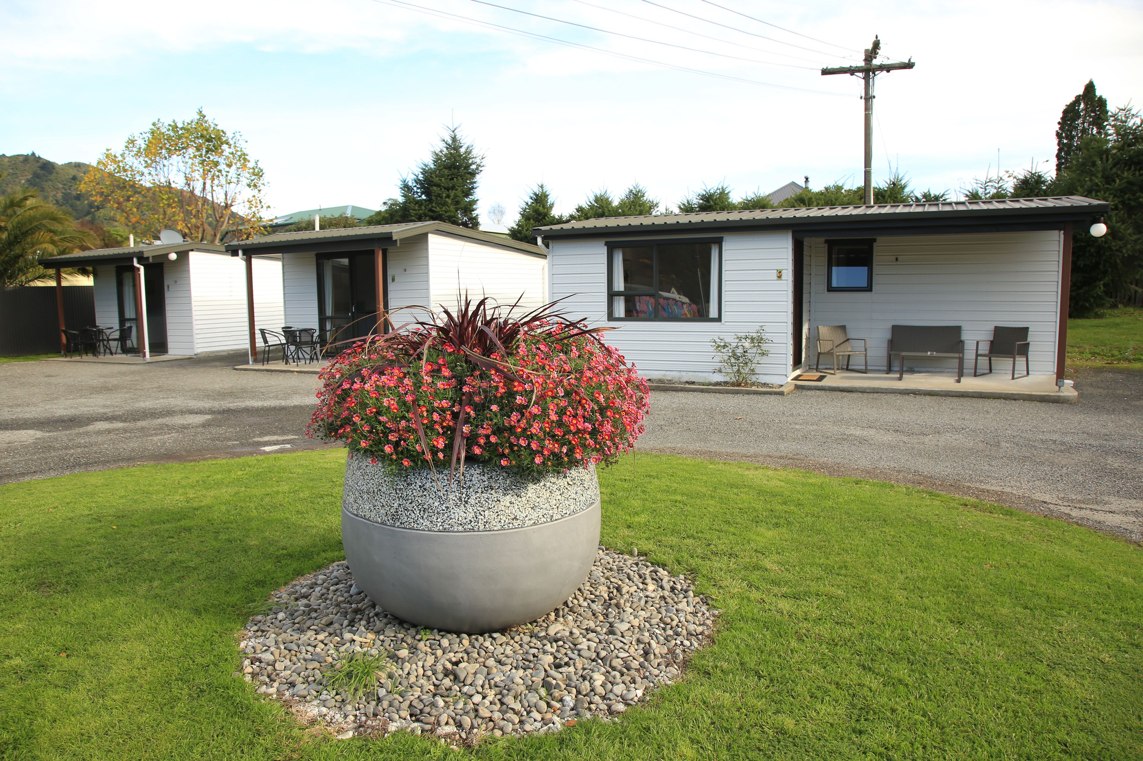 A motel with a large planter of red flowers in front, a gravel driveway, and a grassy area.