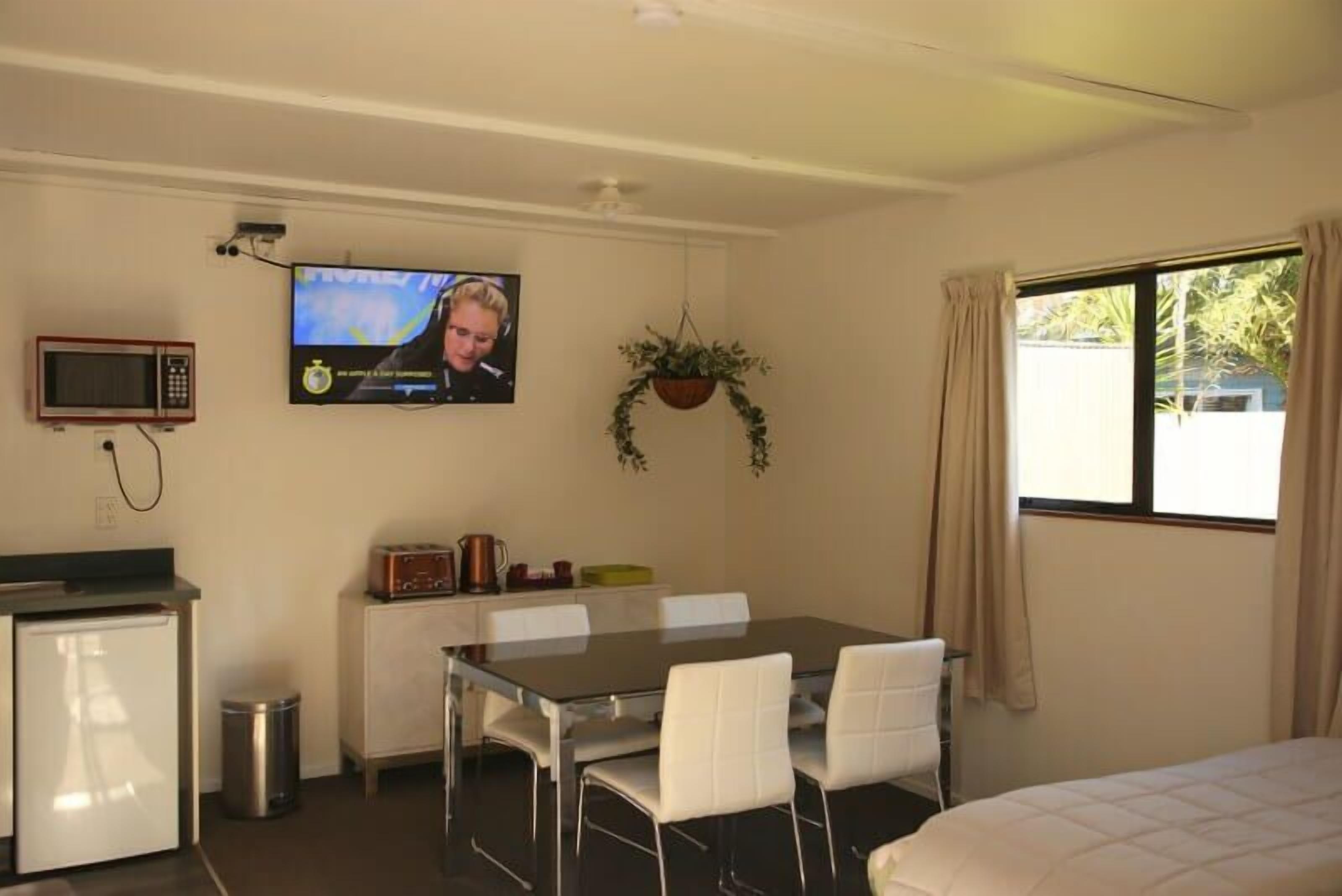 A compact hotel room with a kitchenette, a dining area, a bed, a TV, and a window with curtains.