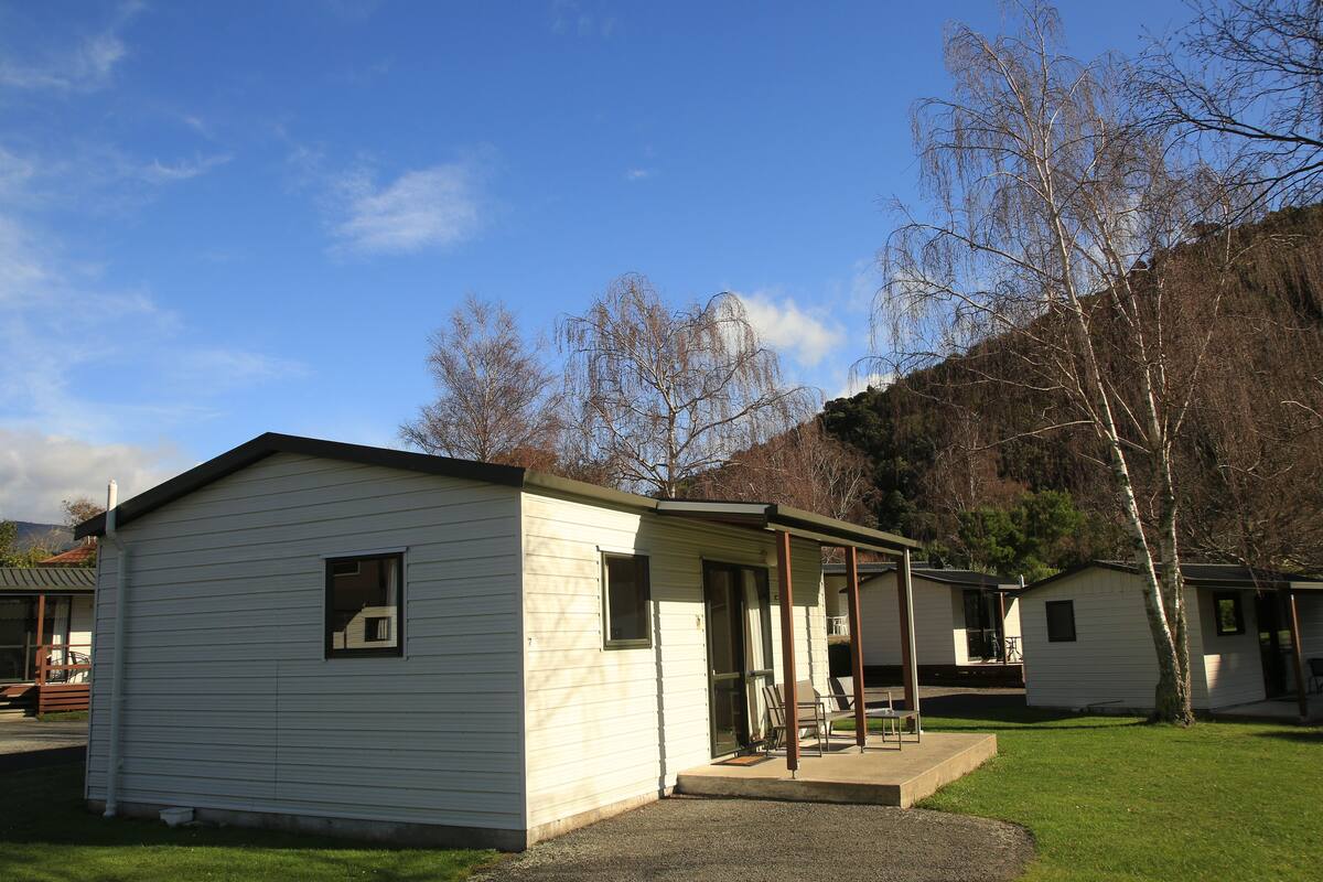 Une rangée de maisons mobiles de plain-pied, dotées d’une véranda couverte, est installée dans un espace verdoyant, avec des arbres et des collines en arrière-plan.