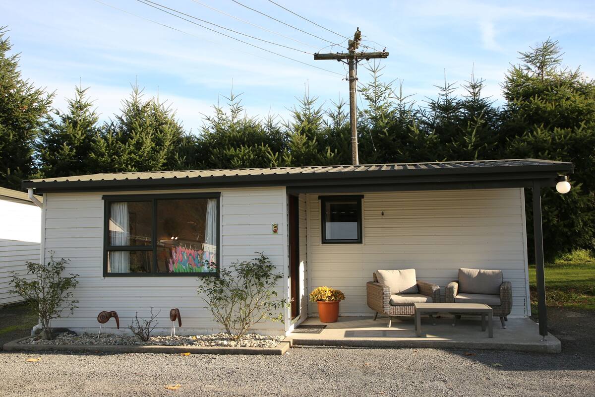 Une petite maison mobile d’un seul étage, dotée d’une véranda, d’une fenêtre et d’un petit jardin agrémenté de plantes et d’objets décoratifs.