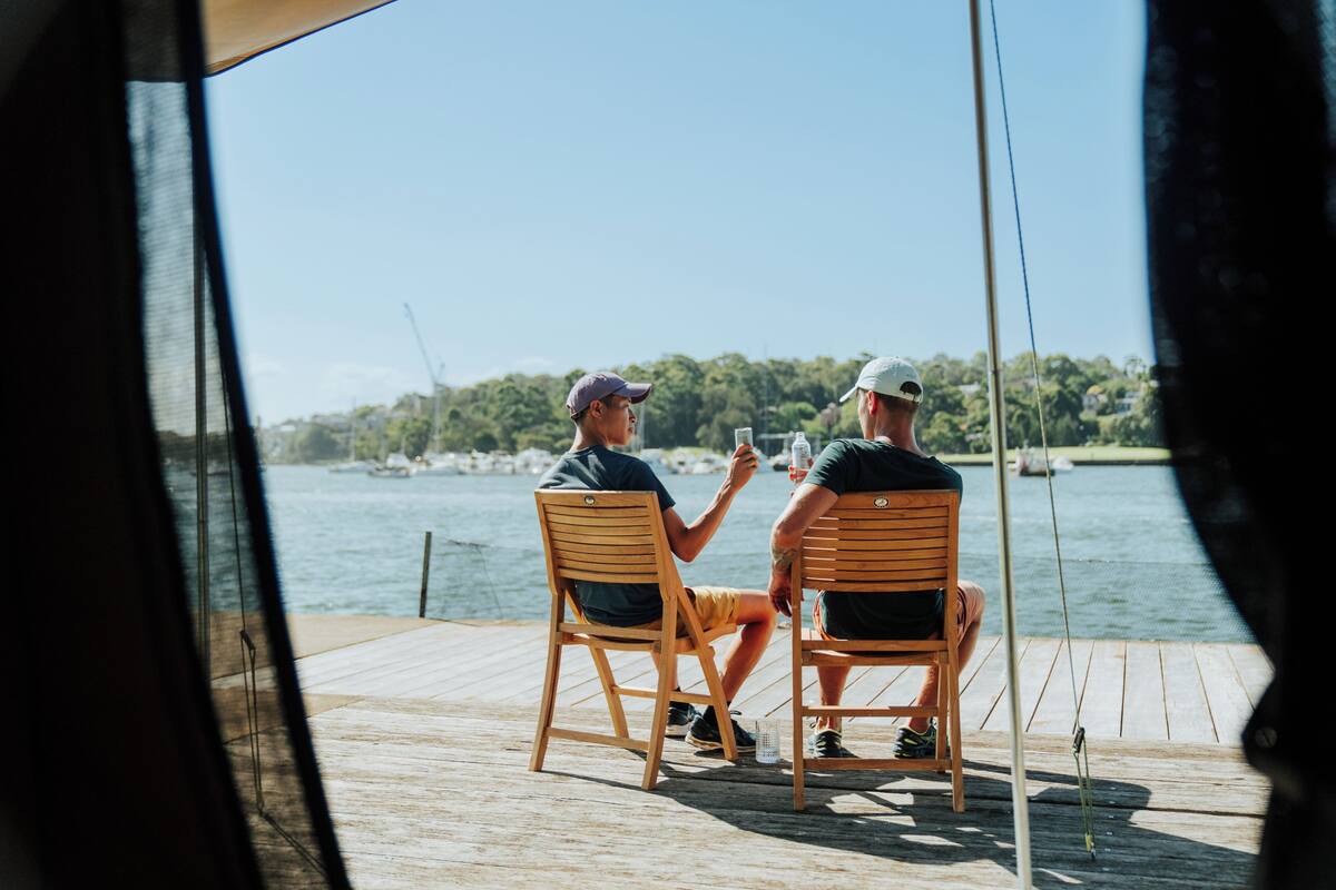 Two people sitting on wooden chairs at a wooden dock, overlooking a body of water with boats and greenery in the background.