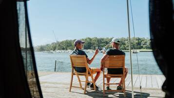 Two people sitting on wooden chairs at a wooden dock, overlooking a body of water with boats and greenery in the background.