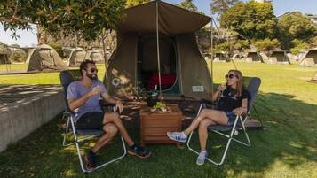 Two people seated at a table under a tent in a camping area with multiple tents in the background.