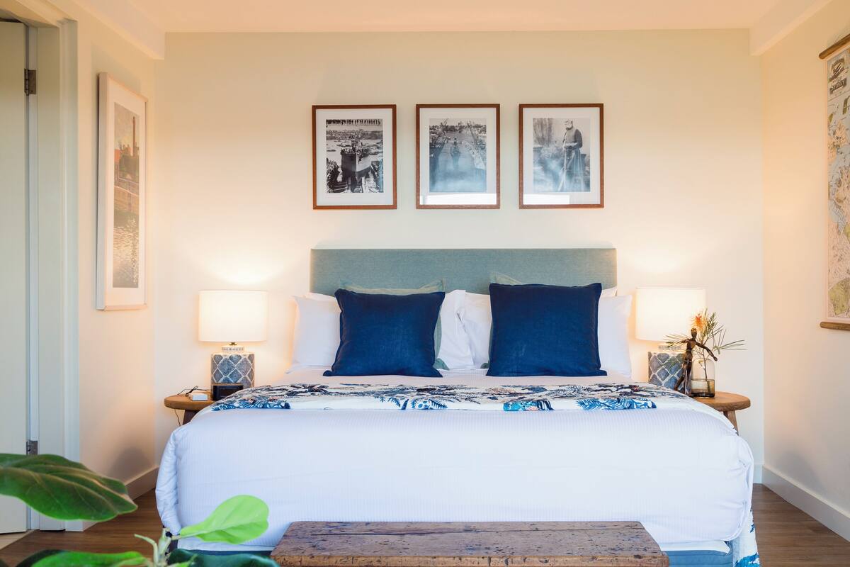 A hotel room with a bed, two bedside tables, a nightstand, a large window, and framed pictures on the wall.