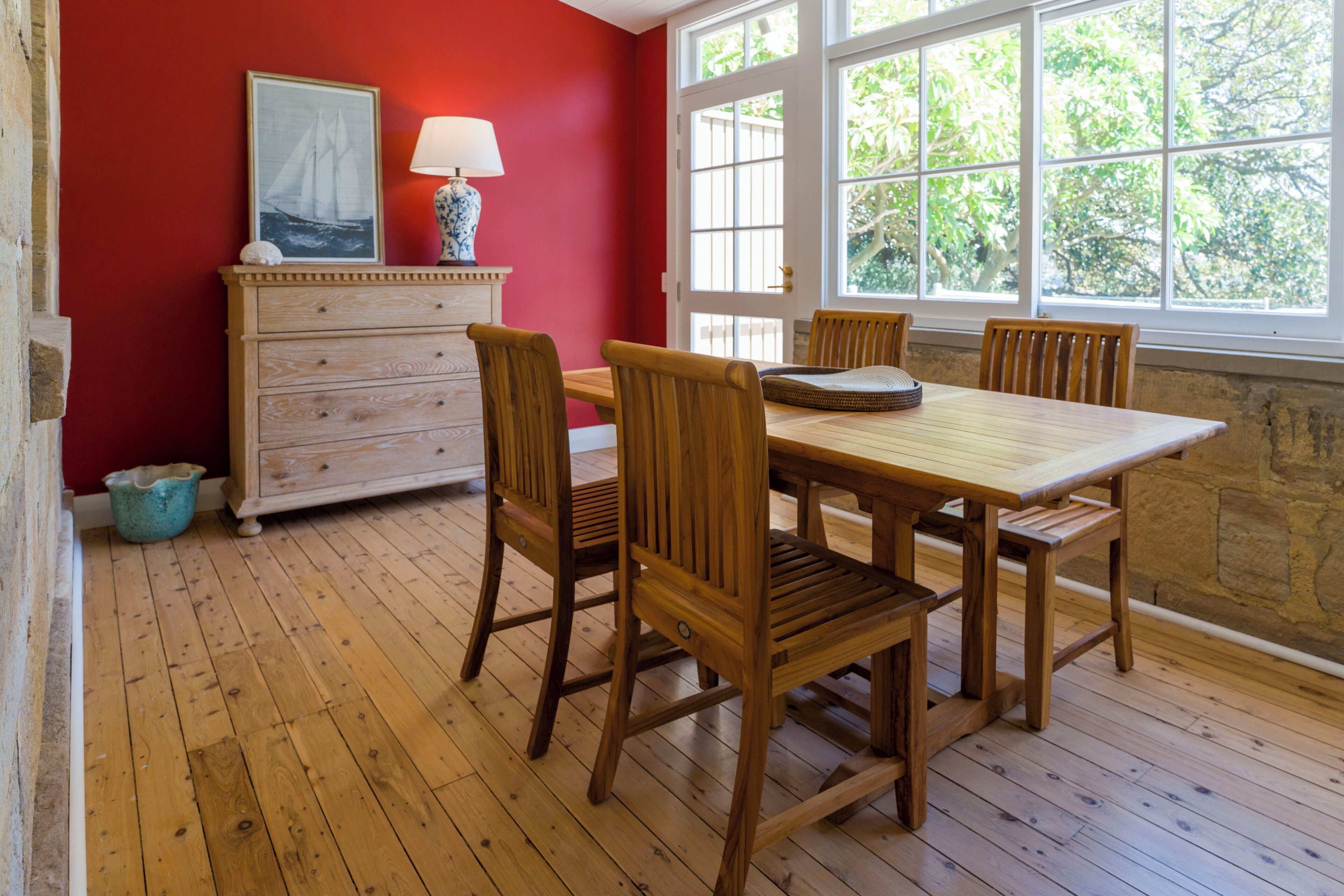 Un coin repas avec une table et des chaises en bois, une commode avec une lampe et une fenêtre donnant sur les arbres.
