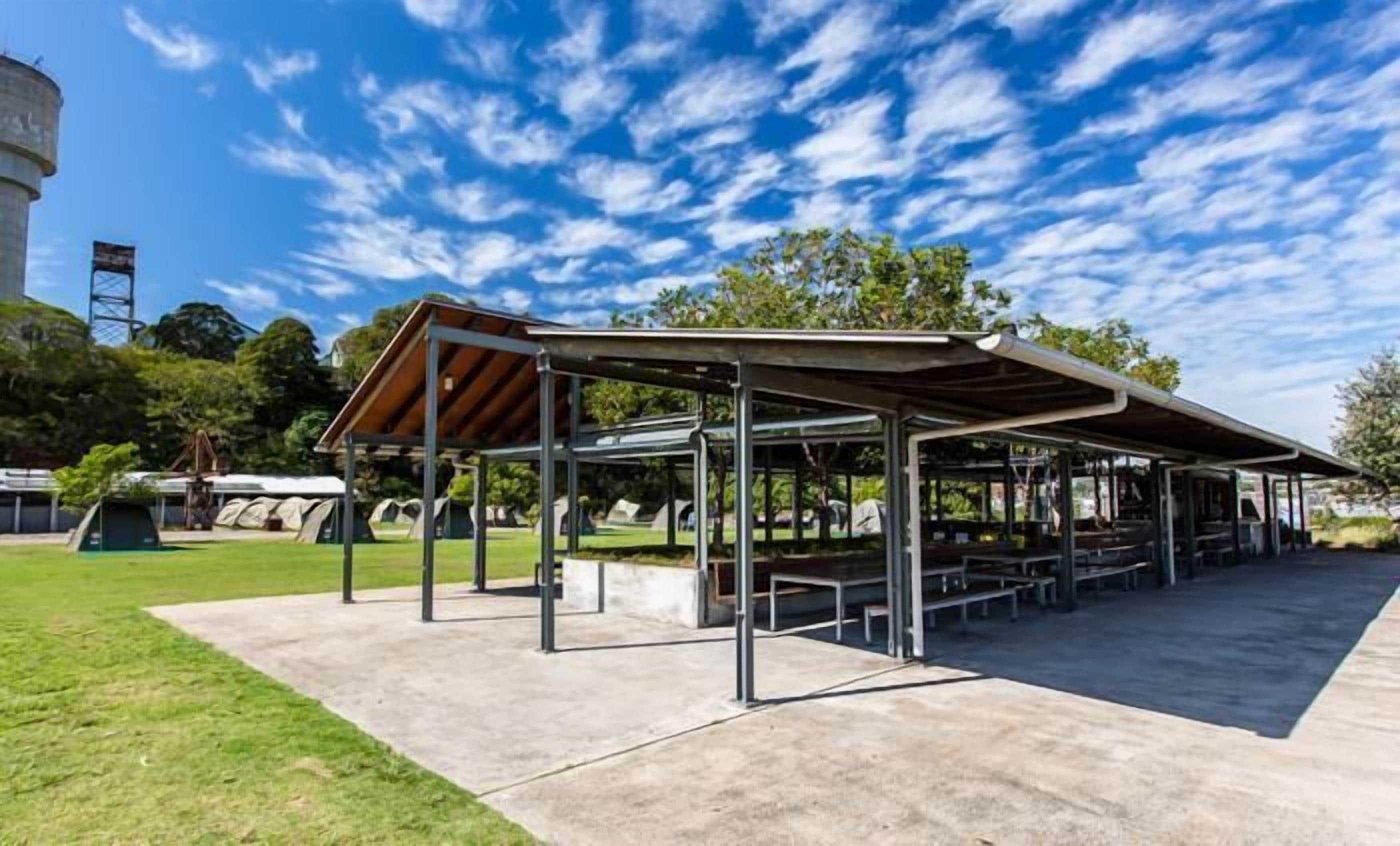 A covered picnic area with metal tables and benches, surrounded by a grassy field and trees.