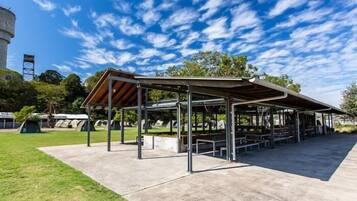 A covered picnic area with metal tables and benches, surrounded by a grassy field and trees.
