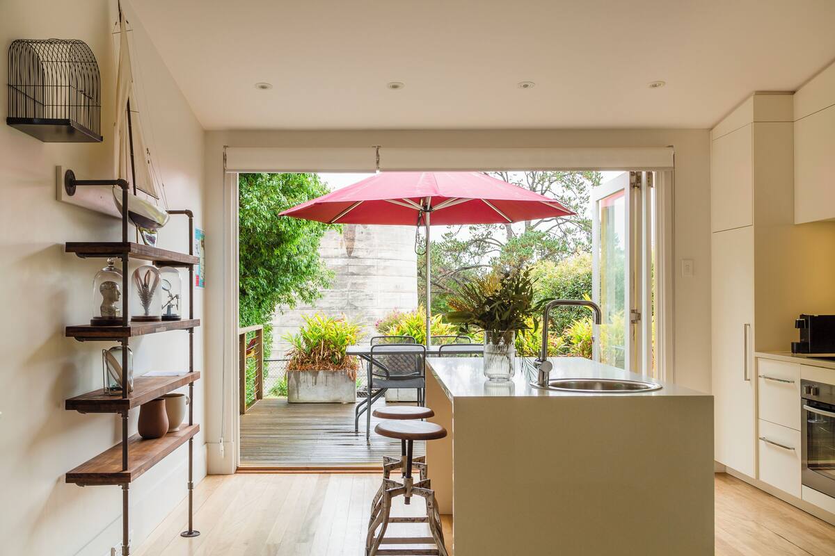A modern kitchen with a central island, a red umbrella, and a view of a garden through glass doors.