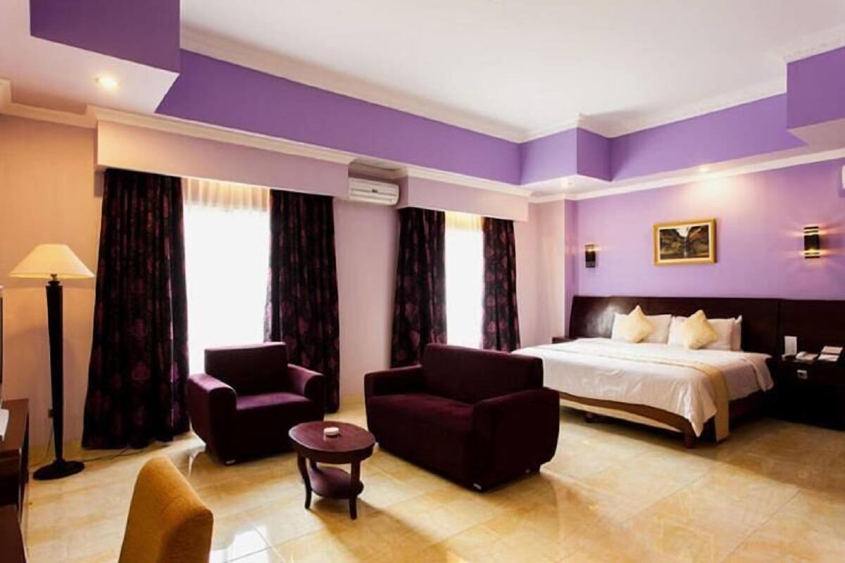 Quarto de hotel com cama, duas poltronas, uma mesinha, um abajur e um quadro na parede.