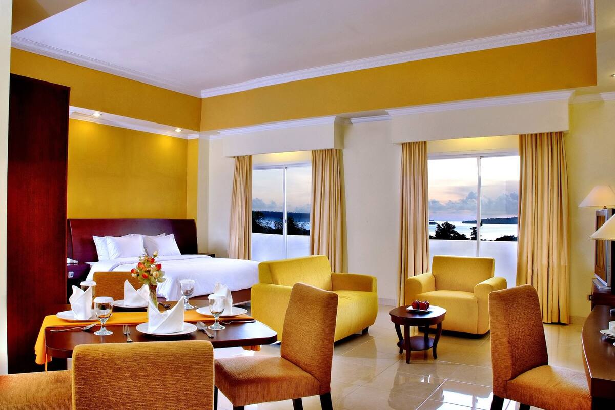 Quarto de hotel com uma cama, duas poltronas amarelas, uma mesa de jantar posta com louça branca e vista para o mar através de grandes janelas.