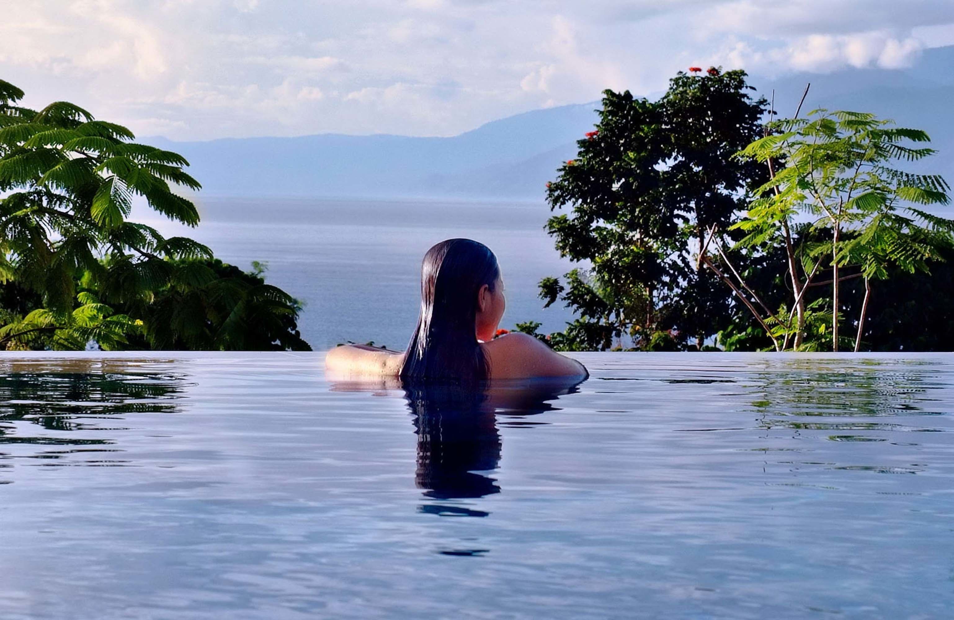 Une personne se trouve dans une piscine à eau claire, avec une vue sur la mer et les montagnes en arrière-plan.