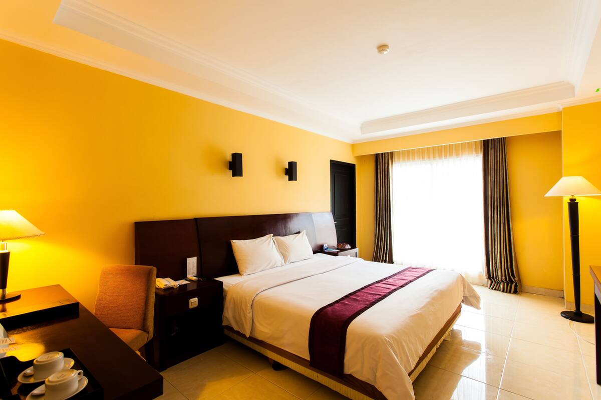 Quarto de hotel com uma cama grande, uma mesa com luminária, uma cadeira e uma mesa de cabeceira com telefone.