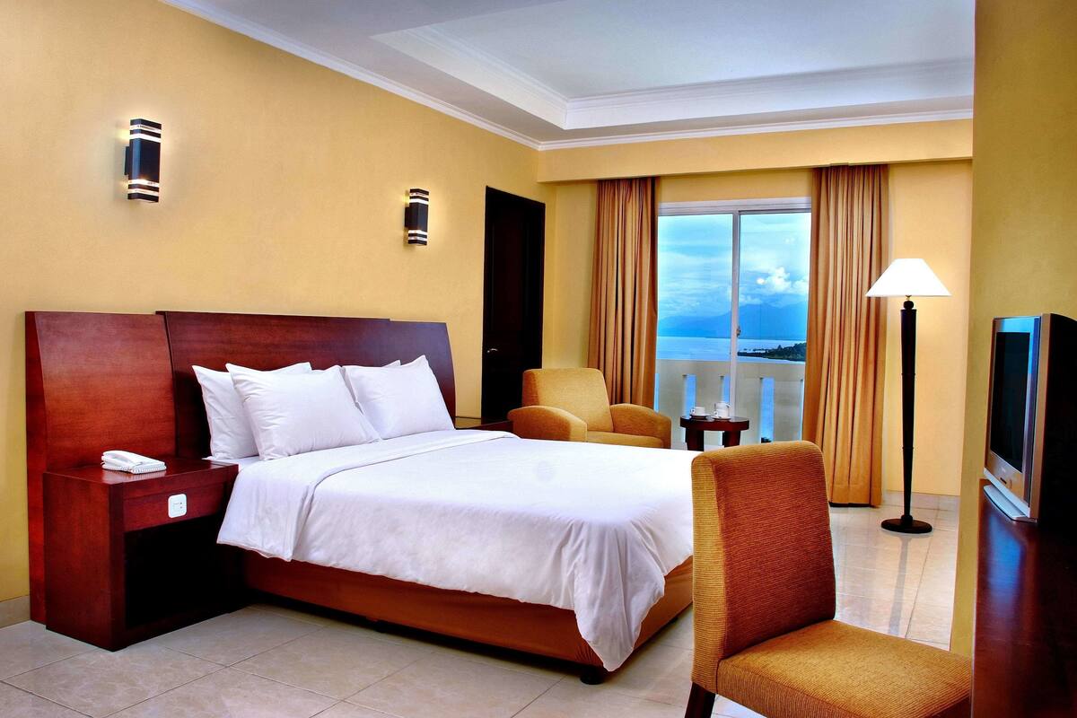 Quarto de hotel com uma cama, uma cadeira, uma televisão, uma mesinha de cabeceira e um abajur.