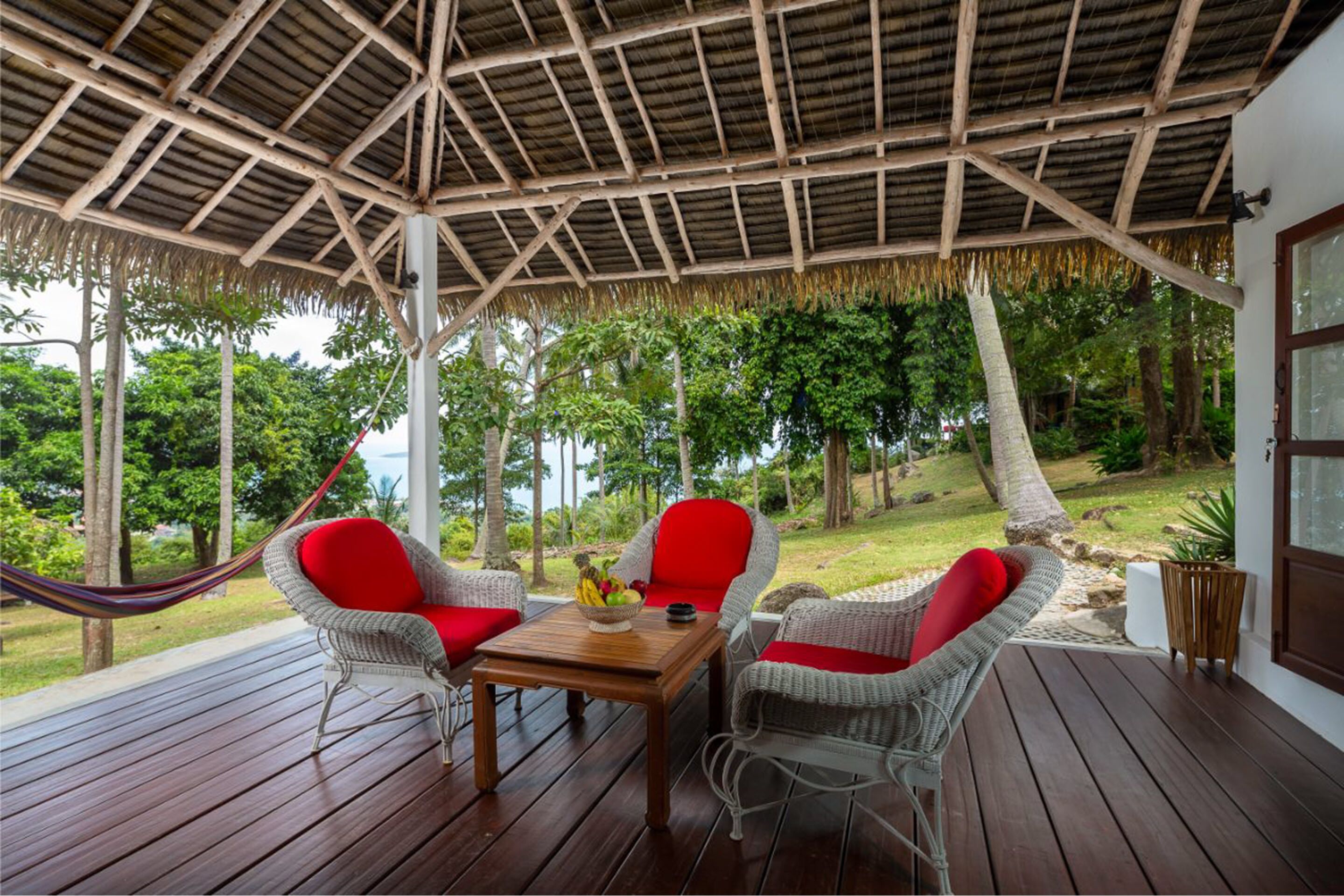 Un espacio de estar al aire libre con techo, con sillas de mimbre y una mesa de madera, con vistas a un jardín exuberante y una masa de agua.