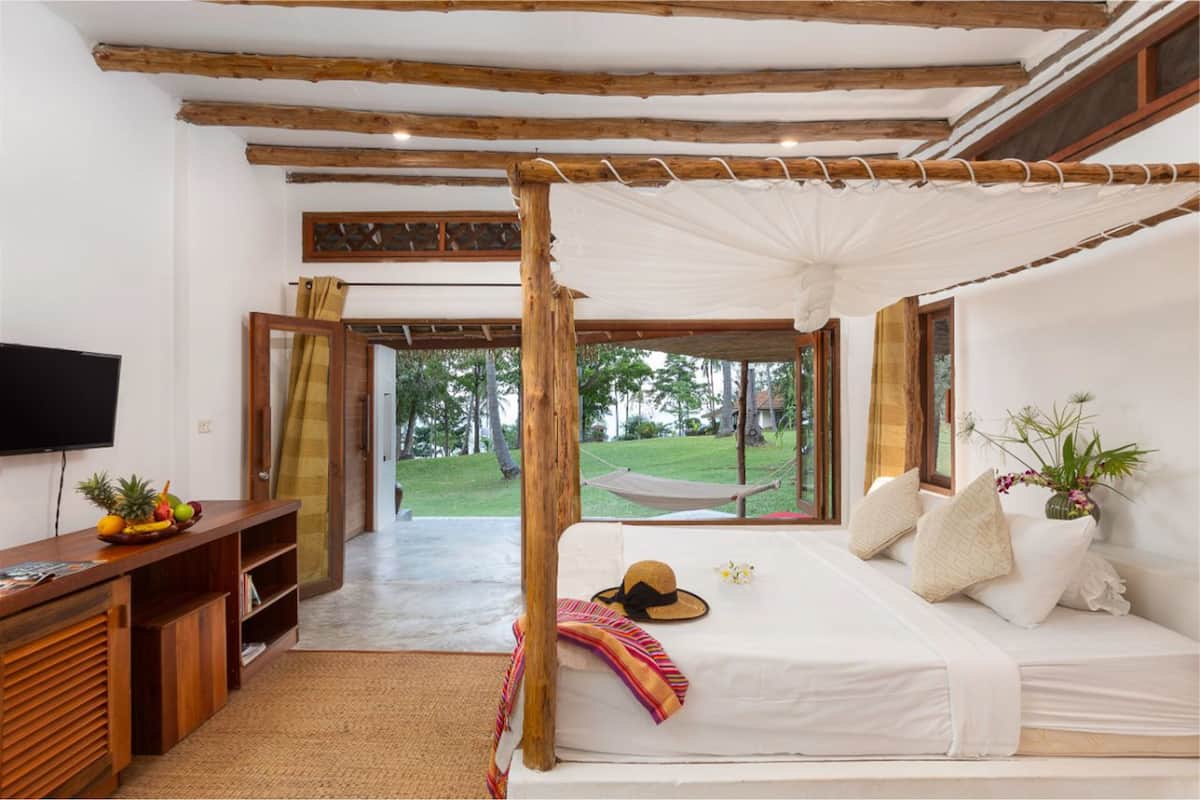 A bedroom with a canopy bed, a TV, a desk with fruit, and a view of the outdoors.