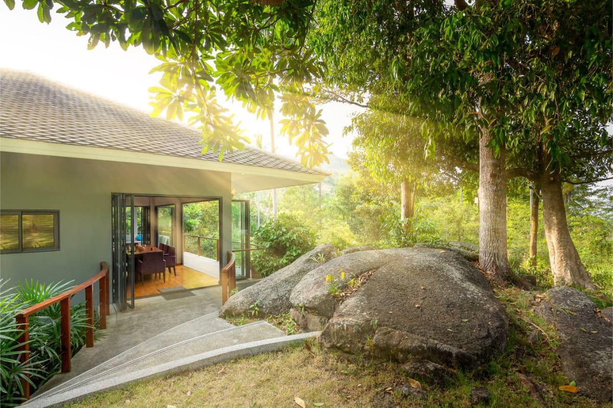A modern house with a glass door entrance, a dining area with a table and chairs, and a large rock in the foreground.