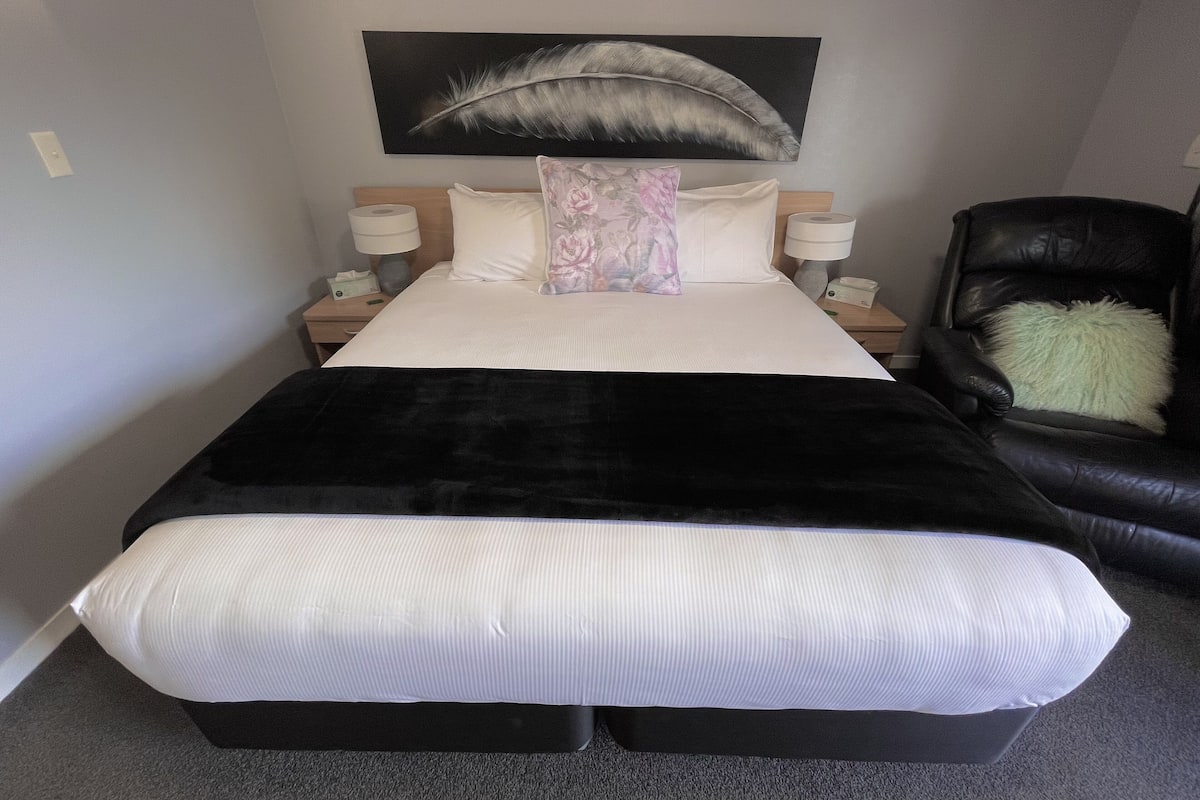 Habitación de hotel con una cama grande, dos mesitas de noche con lámparas, un sillón de cuero negro y una obra de arte con plumas en la pared.