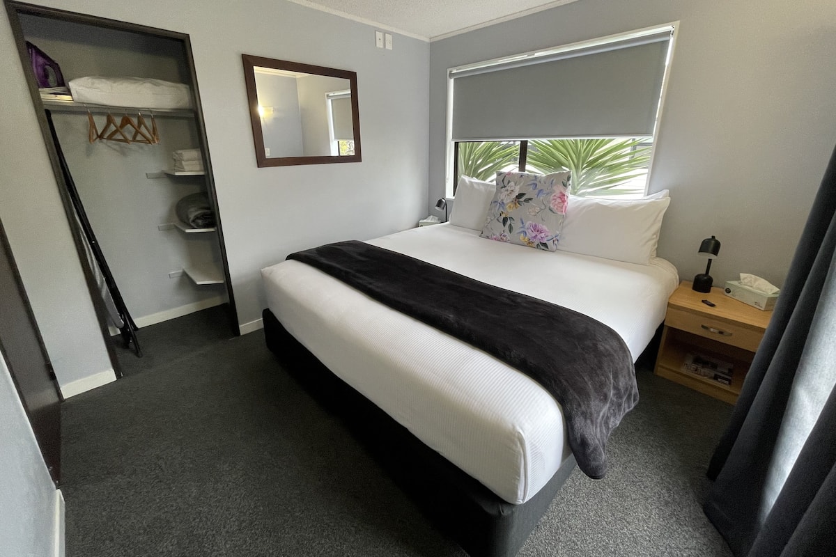 Habitación de hotel ordenada con cama, mesita de noche con lámpara, armario y ventana con vistas a zonas verdes.