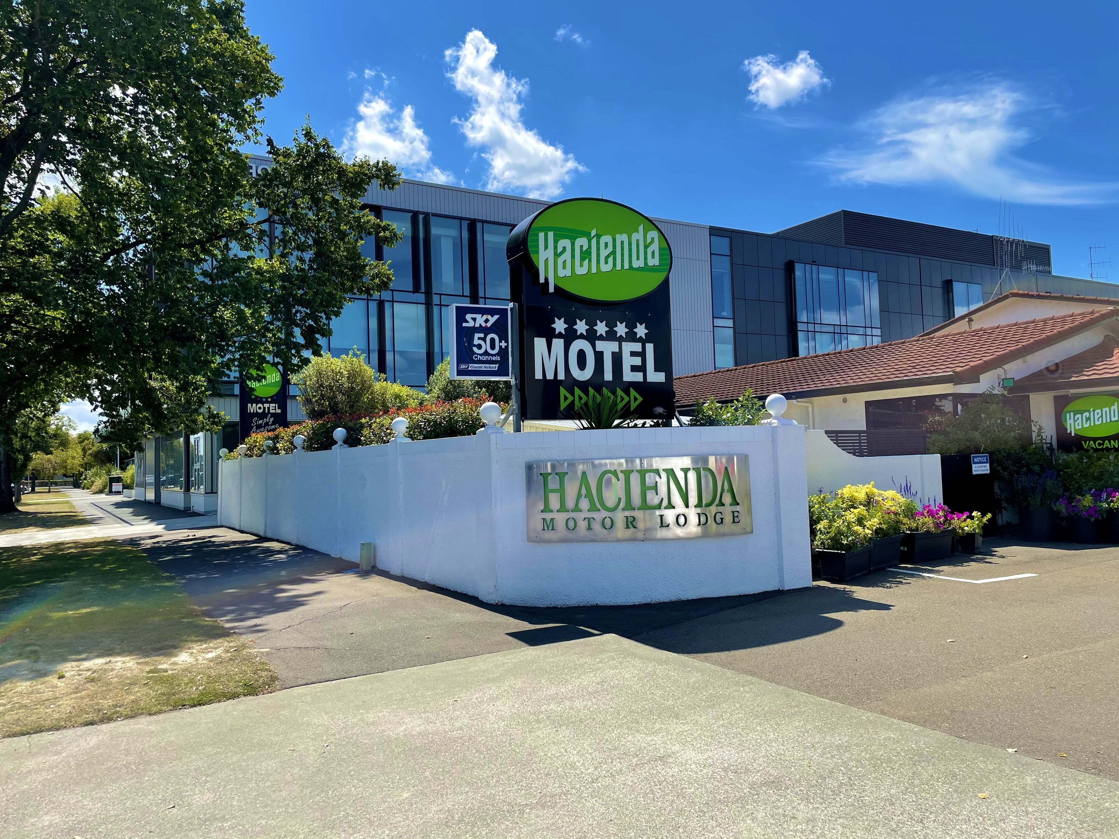 A motel sign with a green and white logo, a Sky 50+ channel sign, and a building with a tiled roof.