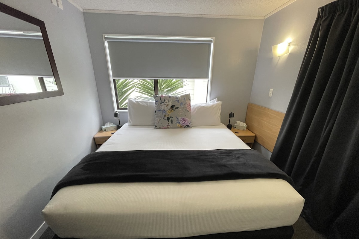 Una habitación de hotel con cama, mesita de noche, lámpara, ventana y vistas a palmeras, además de cortina.