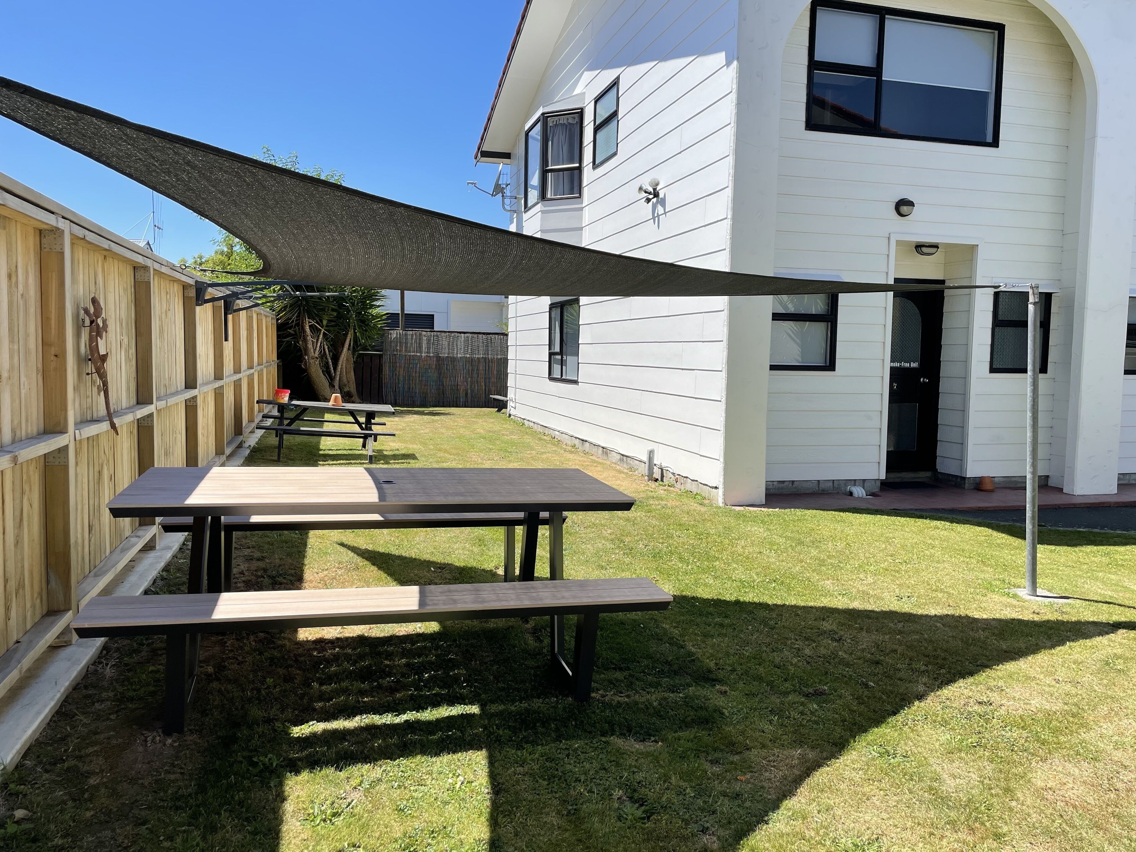 Un jardin avec des tables de pique-nique, une clôture en bois et une maison aux murs recouverts de bardage blanc.