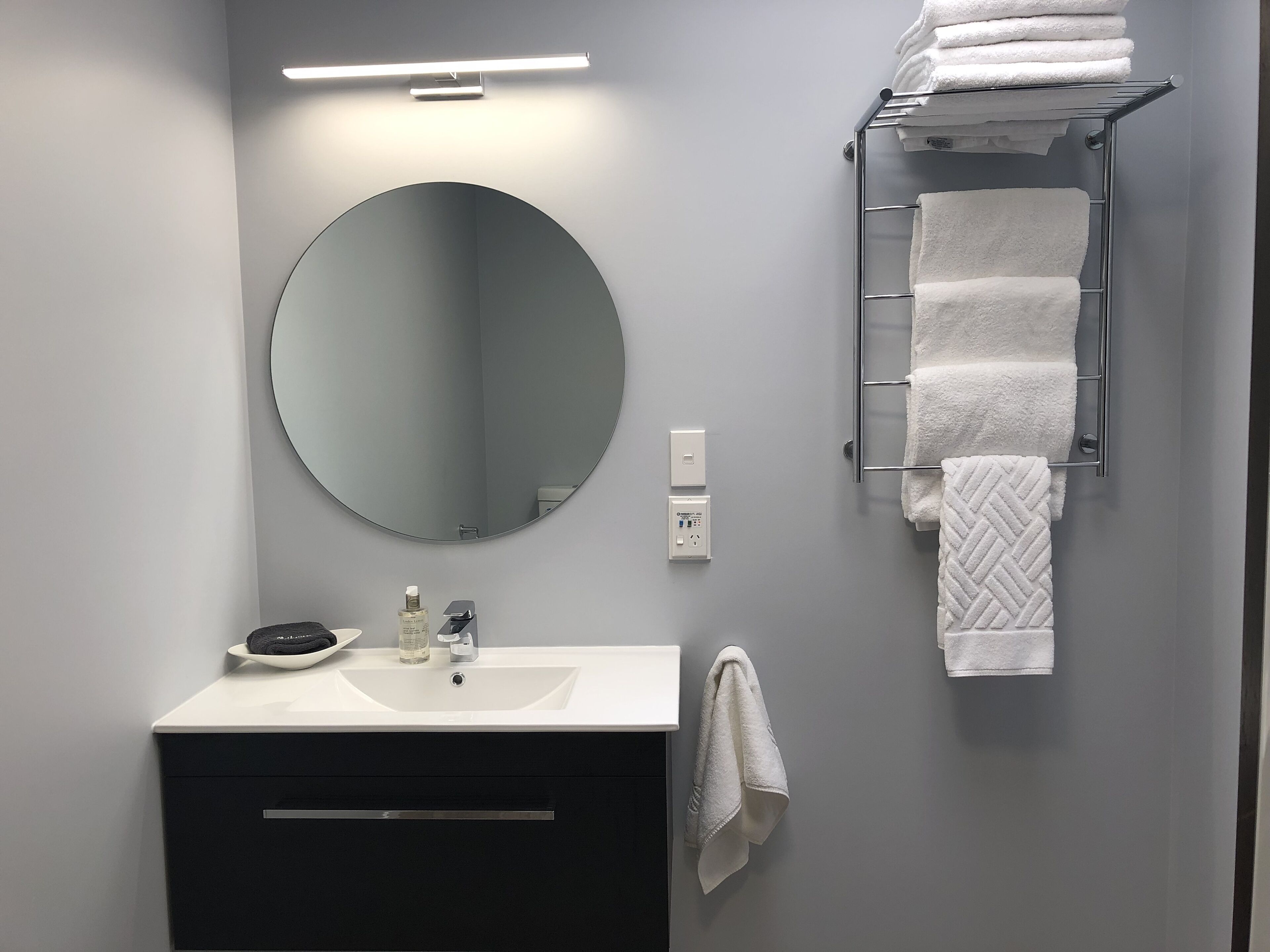 Une salle de bain moderne avec un miroir rond, un lavabo blanc, un porte-serviettes et un luminaire.