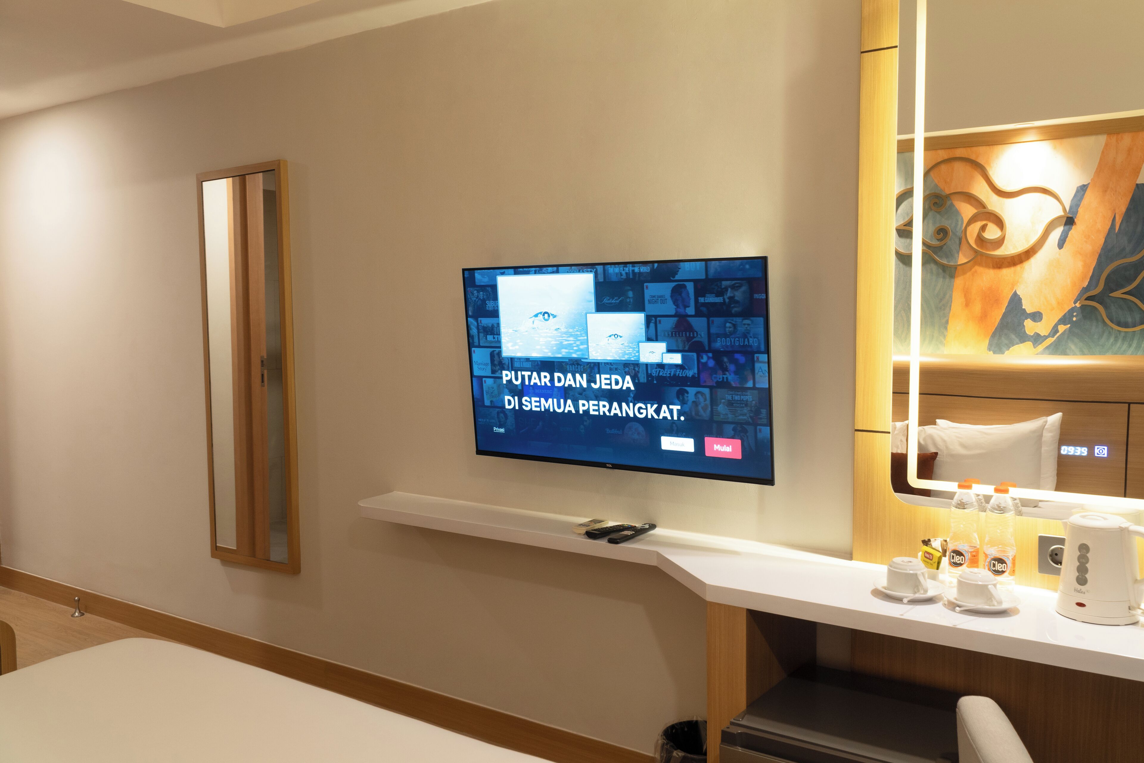 A hotel room with a flat-screen TV mounted on the wall, a wooden side table with a coffee maker, cups, and a bottle of water.