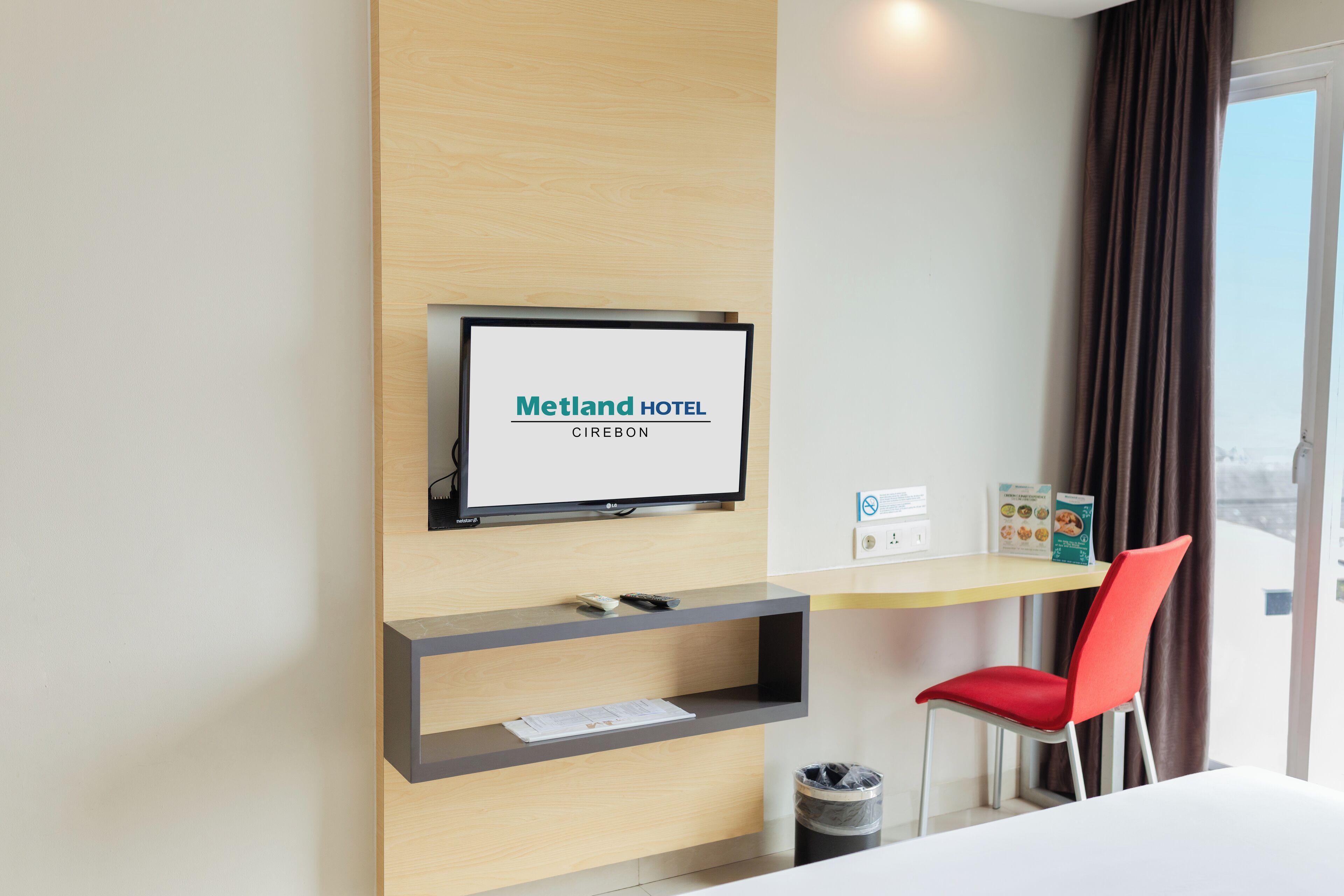 Une chambre d’hôtel équipée d’un téléviseur mural affichant le nom « Metland Hotel Cirebon », d’un bureau avec une chaise et offrant une vue sur la ville par une fenêtre.