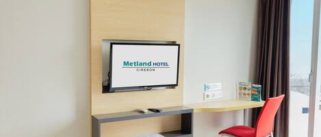 Une chambre d’hôtel équipée d’un téléviseur mural affichant le nom « Metland Hotel Cirebon », d’un bureau avec une chaise et offrant une vue sur la ville par une fenêtre.