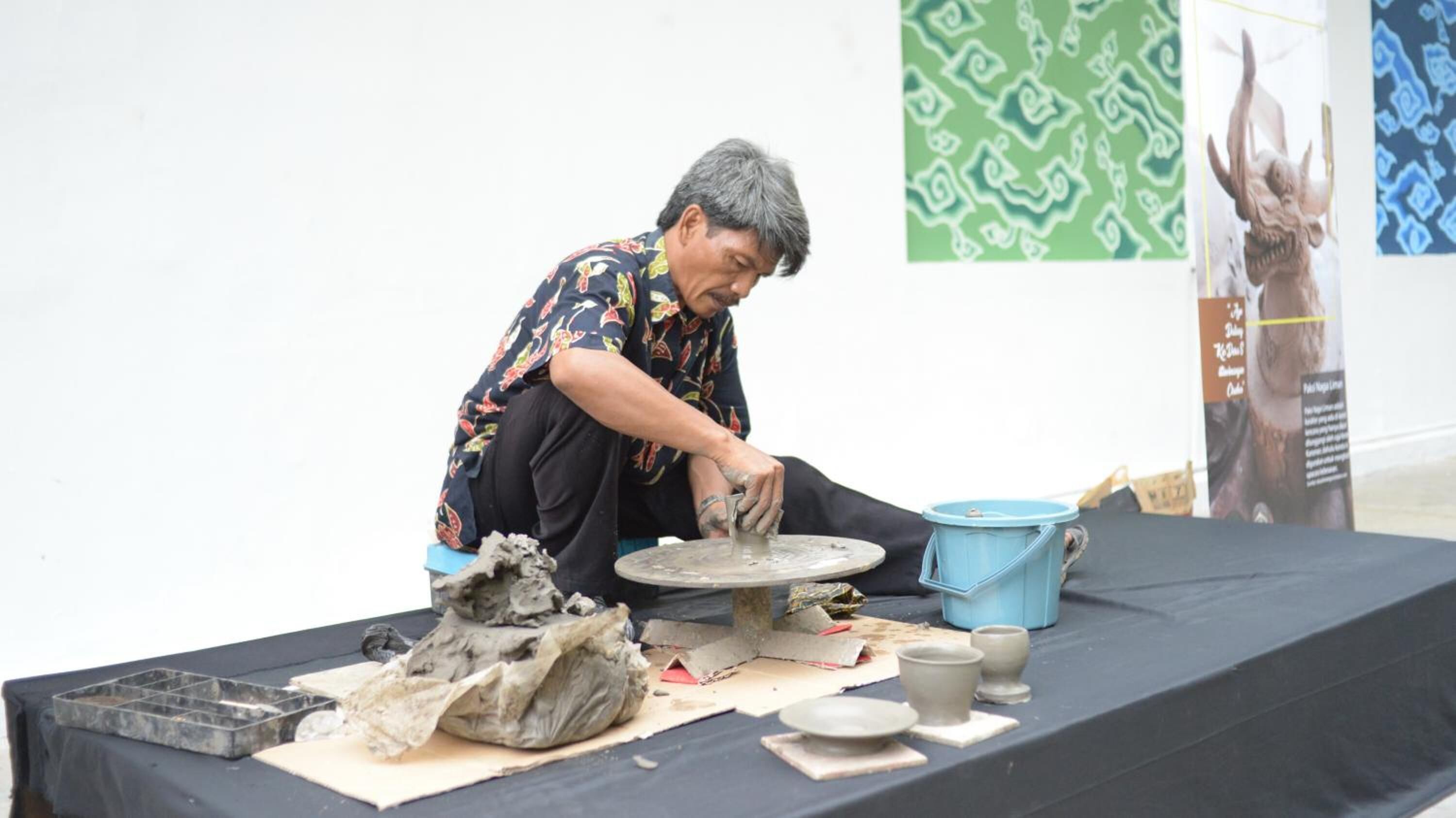 A person working on pottery on a table with a blue bucket, a ceramic wheel, and various pottery pieces.