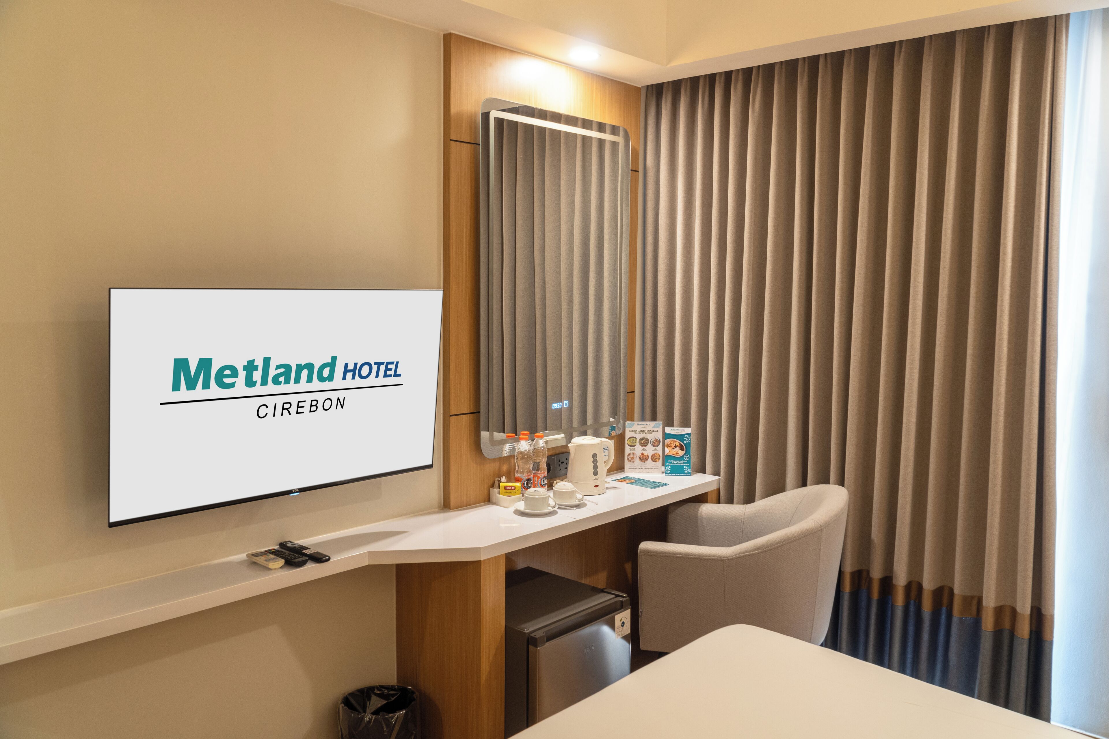 Habitación de hotel con televisor de pantalla plana mostrando 'Metland Hotel Cirebon', un escritorio con cafetera, un cubo de basura y una silla.