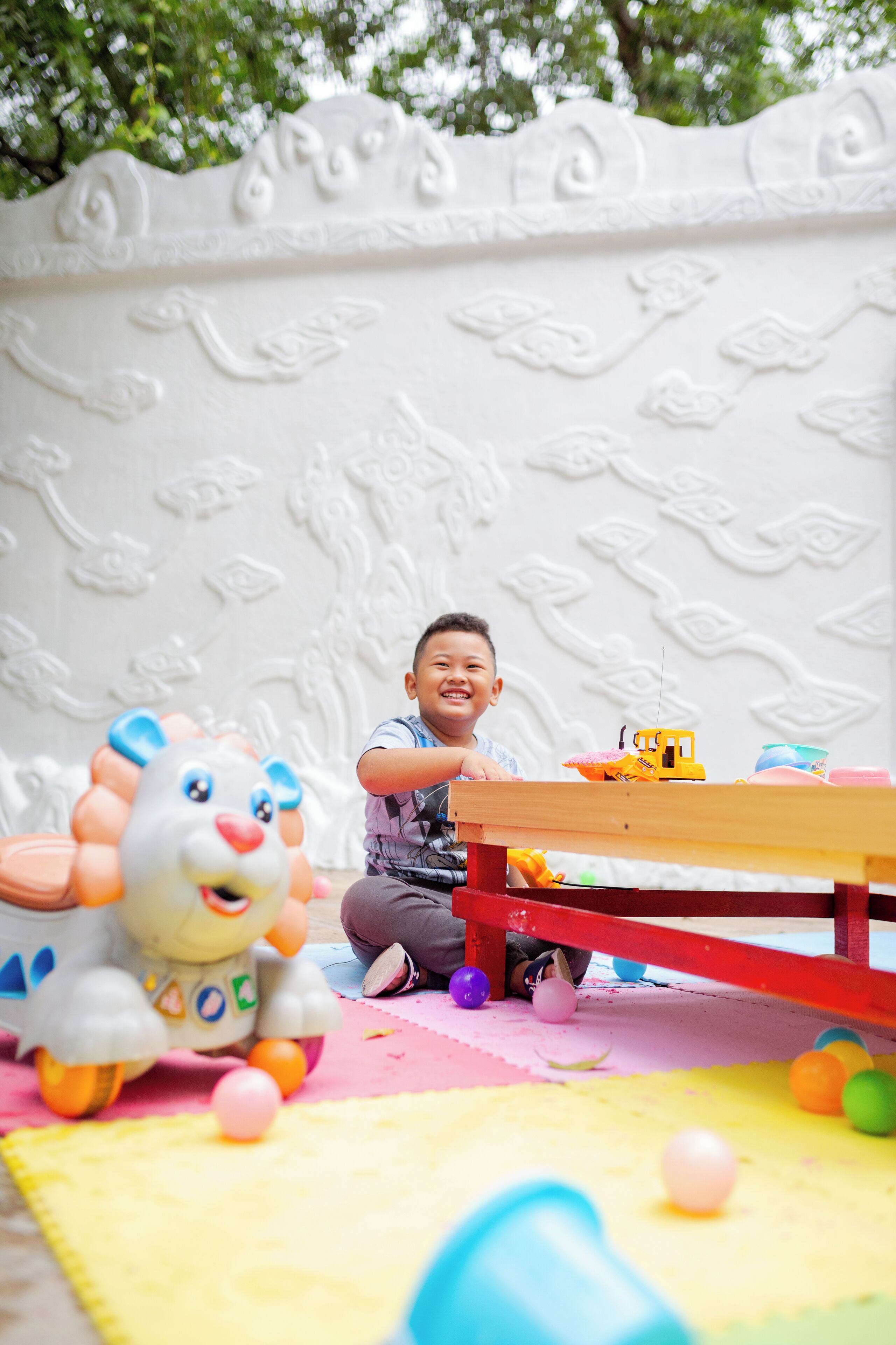A child playing with toys on a colorful play mat with a white wall featuring floral carvings in the background.